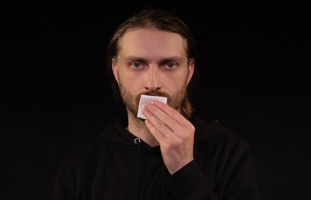 A plainly-dressed man looks towards the camera while kissing a folded piece of paper